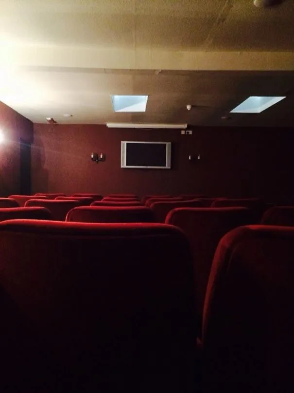 Μικρή αίθουσα προβολής σε ξενοδοχείο με κόκκινα καθίσματα και μια πολύ μικρή τηλεόραση στον τοίχο.