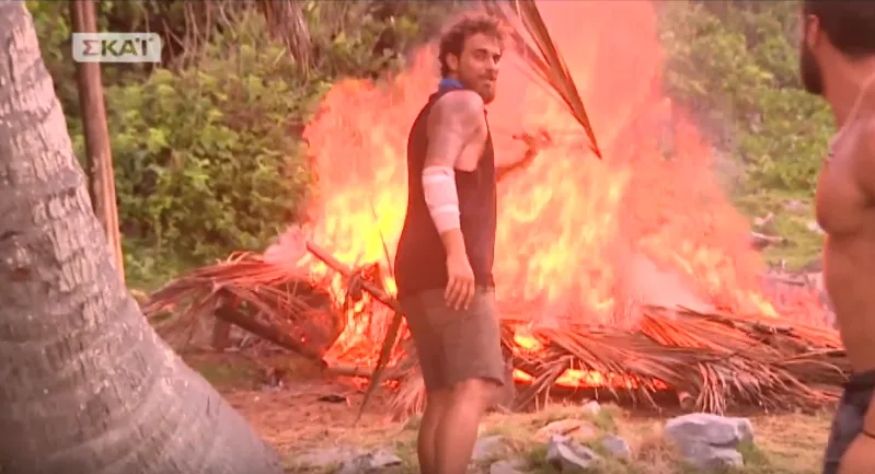 Παίκτης του Survivor μπροστά από μια μεγάλη φωτιά στην παραλία.