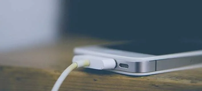 Κοντινό πλάνο ενός iPhone που φορτίζει με καλώδιο Lightning πάνω σε ξύλινη επιφάνεια.