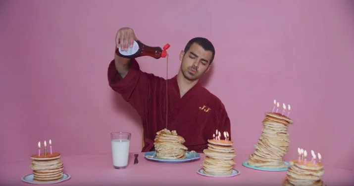 Ο Joe Jonas με μπορντό ρόμπα ρίχνει σιρόπι σε στοίβες από pancakes με κεράκια σε ροζ φόντο.