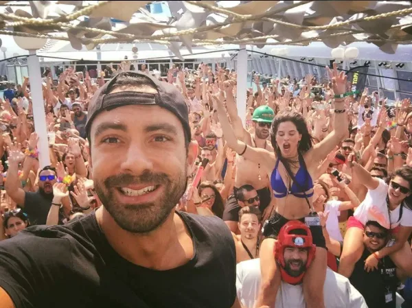 Ο Σάκης Τανιμανίδης βγάζει selfie σε ένα κατάστρωμα πλοίου γεμάτο κόσμο που διασκεδάζει.