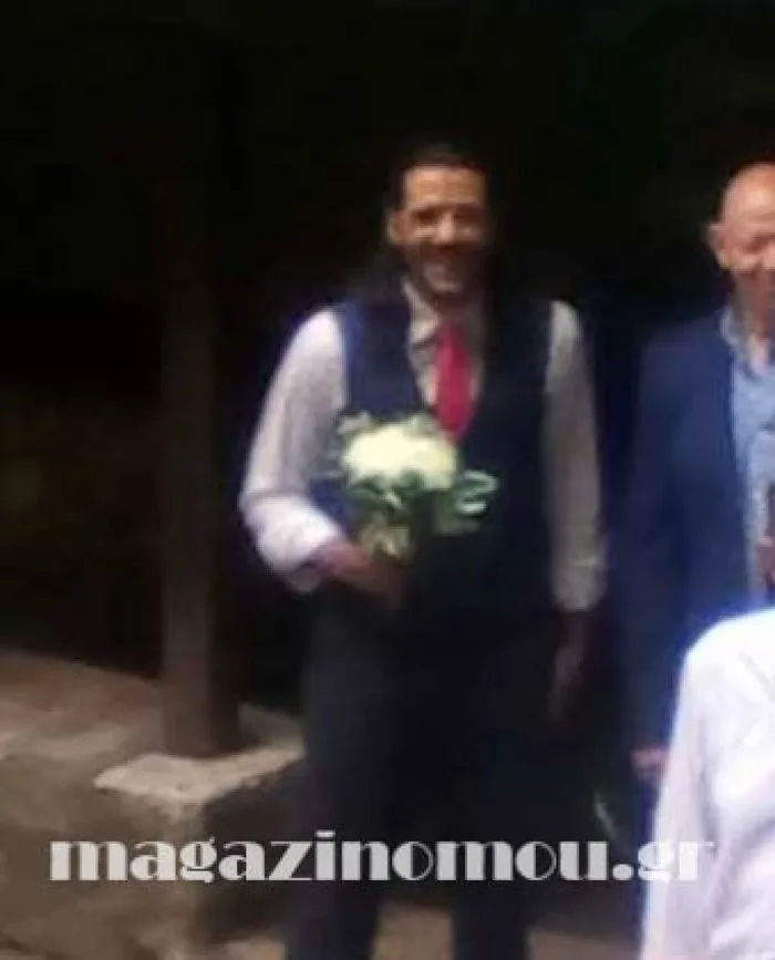 Άνδρας με γιλέκο και κόκκινη γραβάτα κρατάει ανθοδέσμη σε εξωτερικό χώρο.