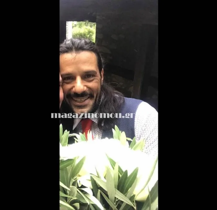 Έλληνας ηθοποιός με κοστούμι και κόκκινη γραβάτα κρατάει λευκά λουλούδια.