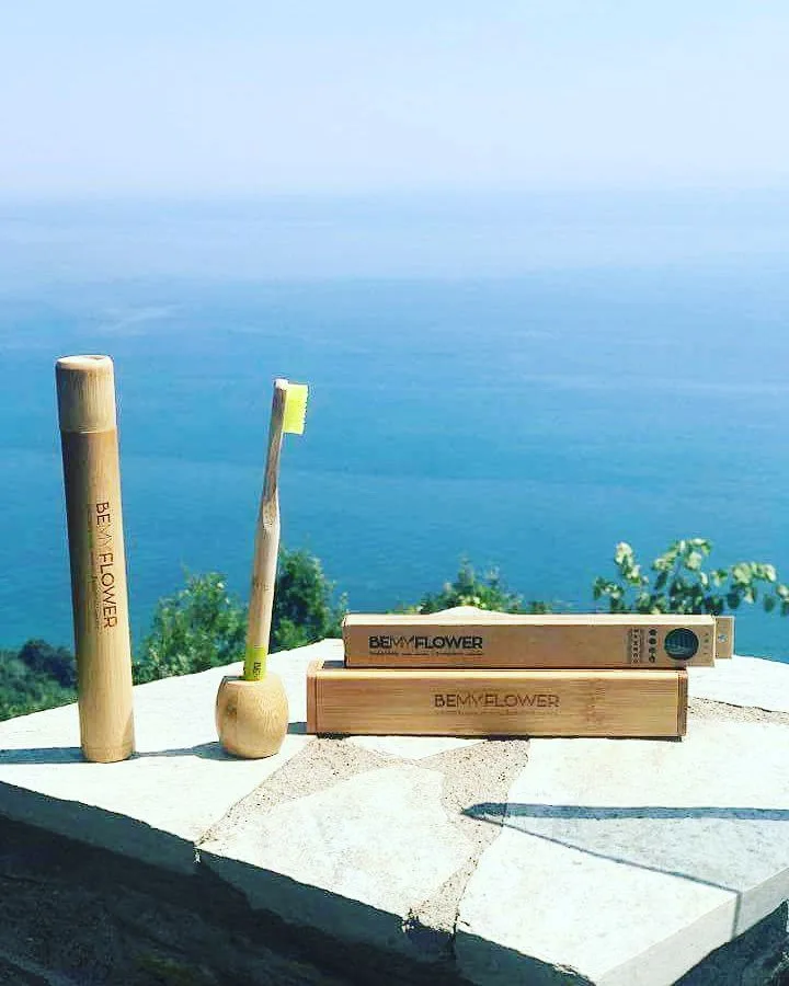 Ξύλινες οδοντόβουρτσες και θήκες BeMyFlower σε πέτρινο πεζούλι με φόντο τη θάλασσα.