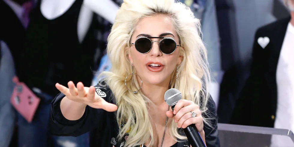 Η Lady Gaga με ξανθά μαλλιά και στρογγυλά γυαλιά ηλίου κρατάει μικρόφωνο.