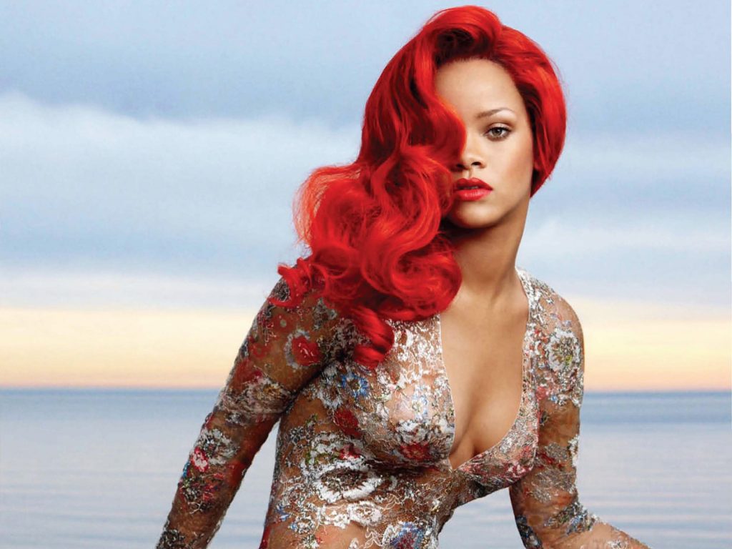 Η Rihanna με έντονα κόκκινα μαλλιά και δαντελωτό φόρεμα σε φόντο θάλασσας.