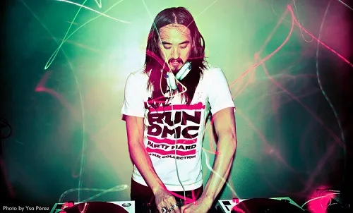 DJ με μακριά μαλλιά και λευκό t-shirt παίζει μουσική σε πάρτι με εφέ φωτισμού.