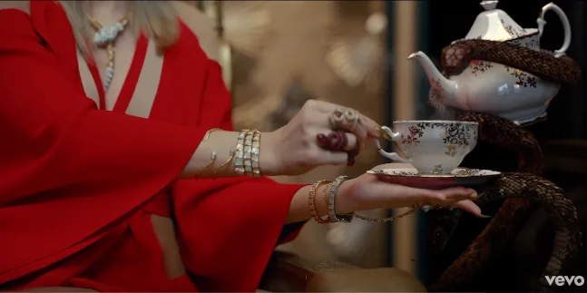 Η Taylor Swift με κόκκινο ένδυμα κρατάει ένα φλιτζάνι τσάι δίπλα σε μια τσαγιέρα με φίδι.