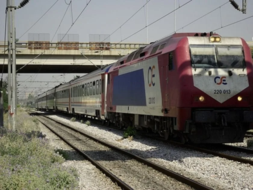 Επιβατική αμαξοστοιχία του ΟΣΕ κινείται σε σιδηροδρομικές γραμμές κάτω από γέφυρα.