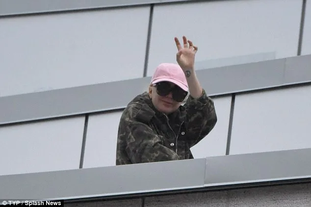 Η Lady Gaga με ροζ καπέλο και καμουφλάζ τζάκετ χαιρετά από μπαλκόνι.