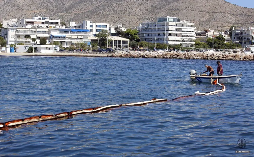 Πλωτό φράγμα στη θάλασσα για τον περιορισμό πετρελαιοκηλίδας με βάρκα και κτίρια στο βάθος.