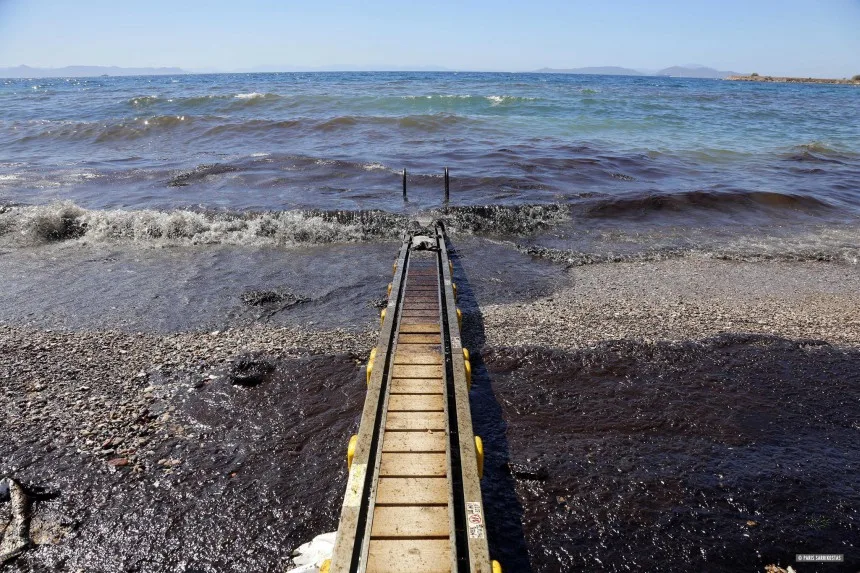 Πετρελαιοκηλίδα σε παραλία της Γλυφάδας με μαύρα νερά και μια ράμπα πρόσβασης στη θάλασσα.