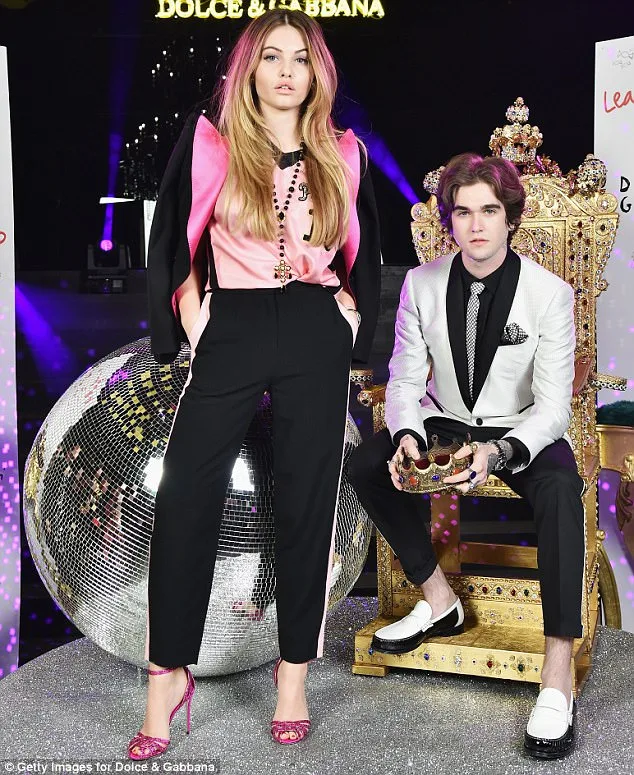 Η Thylane Blondeau με ροζ τοπ και μαύρο παντελόνι δίπλα σε άνδρα που κάθεται σε θρόνο.