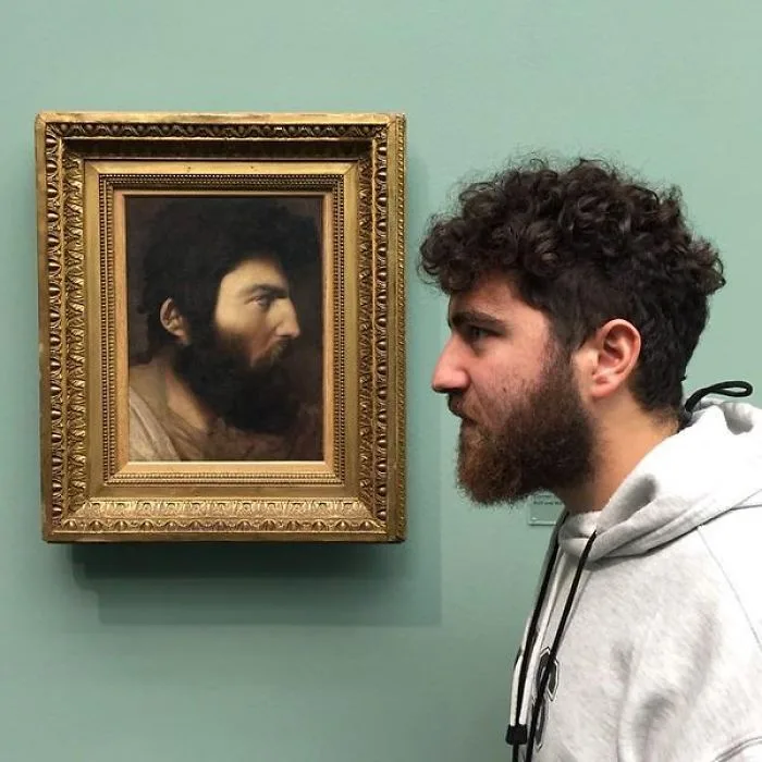 Ένας άνδρας με μούσια και σγουρά μαλλιά δίπλα σε πίνακα που απεικονίζει τον σωσία του.