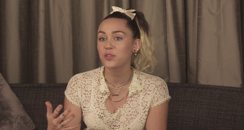 Η Miley Cyrus με ξανθά μαλλιά πιασμένα ψηλά και δαντελωτό μπλουζάκι.