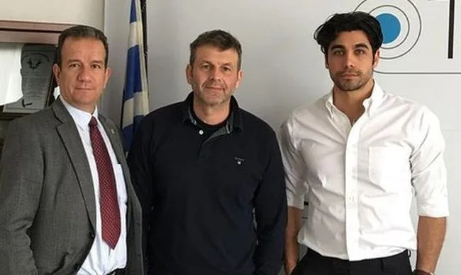 Τρεις άνδρες ποζάρουν σε εσωτερικό χώρο μπροστά από μια ελληνική σημαία.