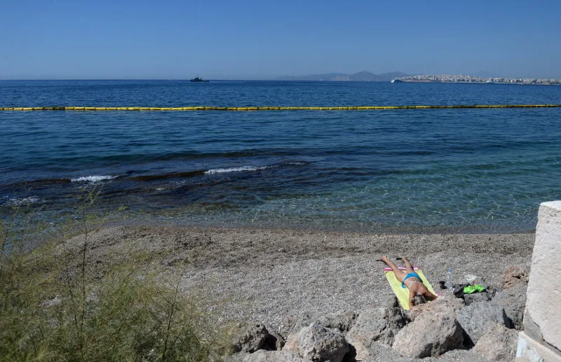 Παραλία με βότσαλα και μια γυναίκα να κάνει ηλιοθεραπεία, ενώ στη θάλασσα διακρίνεται ρύπανση.