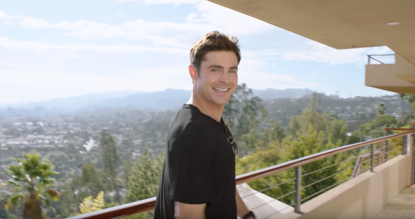 Ο Zac Efron χαμογελά σε ένα μπαλκόνι με θέα την πόλη.