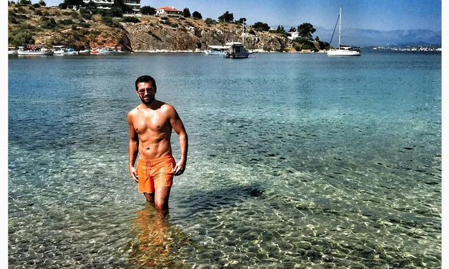 Άνδρας με πορτοκαλί μαγιό στέκεται στα ρηχά νερά μιας παραλίας.