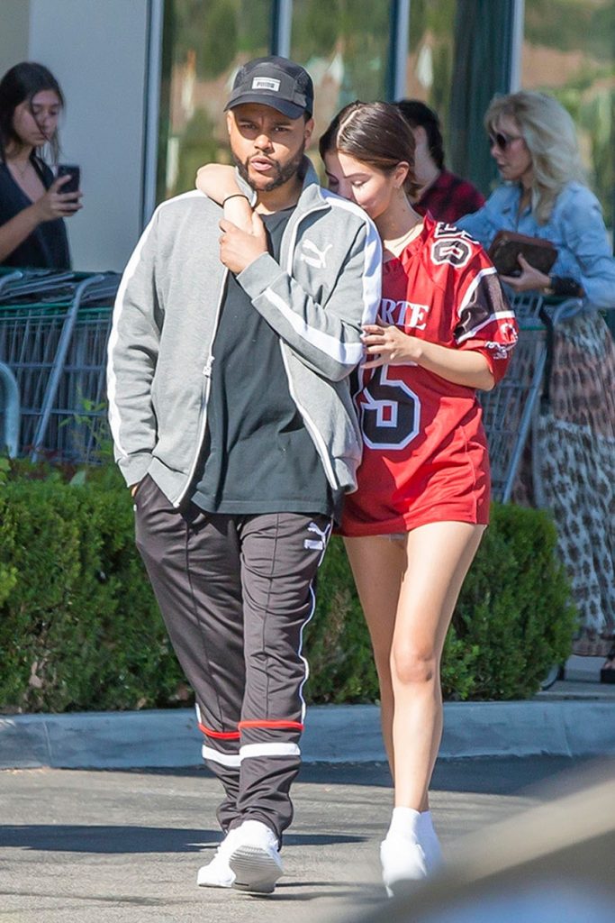 Ο The Weeknd και η Selena Gomez περπατούν αγκαλιασμένοι σε εξωτερικό χώρο.