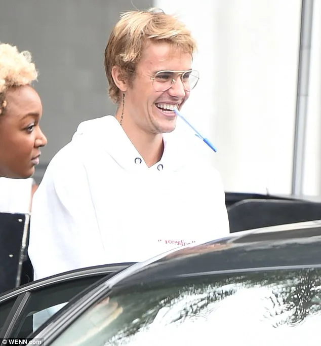 Ο Justin Bieber με λευκό φούτερ και γυαλιά ηλίου κρατώντας ένα στυλό στο στόμα.