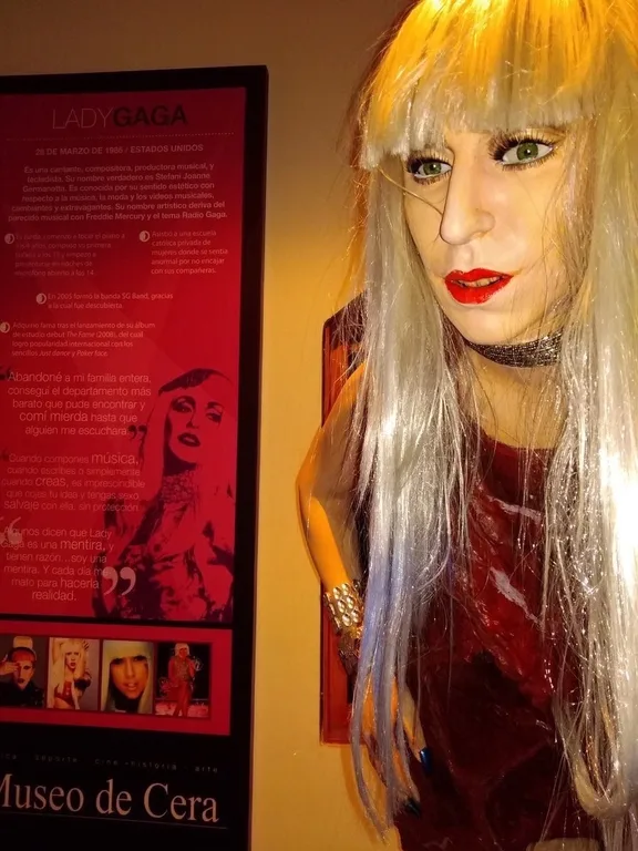 Κέρινο ομοίωμα της Lady Gaga σε μουσείο με ενημερωτικό πίνακα στα αριστερά.