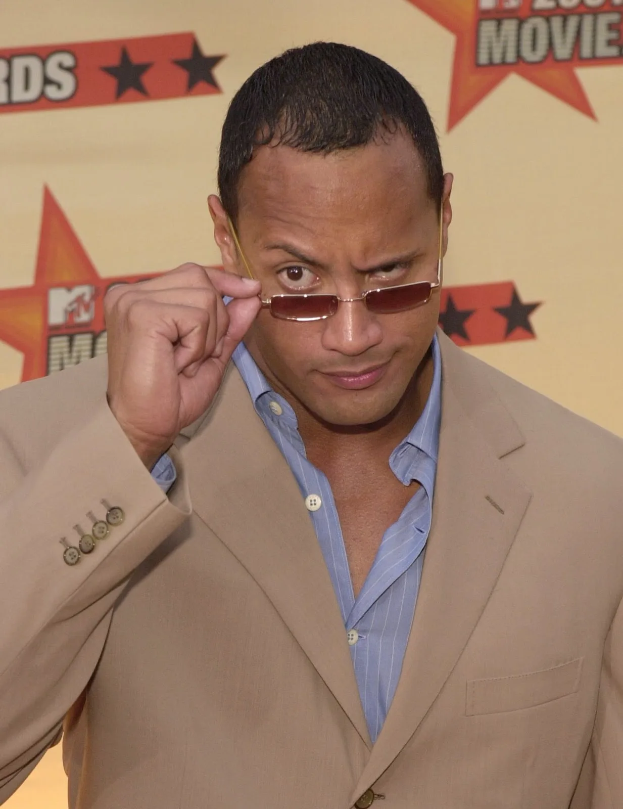 Ο Dwayne Johnson με μικροσκοπικά γυαλιά ηλίου και μπεζ κοστούμι.
