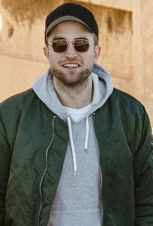 Ο Robert Pattinson με καπέλο, γυαλιά ηλίου, γκρι φούτερ και πράσινο μπουφάν.