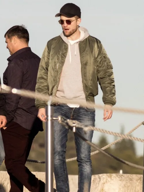 Ο Robert Pattinson με λαδί bomber jacket, γκρι φούτερ και καπέλο σε εξωτερικό χώρο.