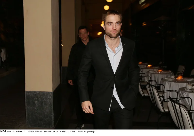 Ο Robert Pattinson με μαύρο κοστούμι και ανοιχτό πουκάμισο σε εξωτερικό χώρο εστιατορίου.