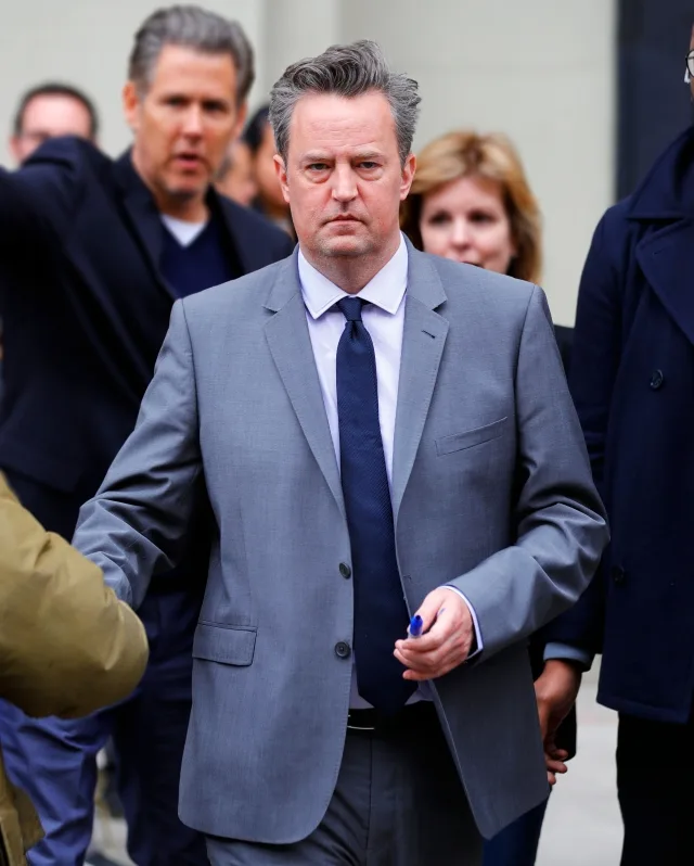 Ο Matthew Perry με γκρι κοστούμι και μπλε γραβάτα σε εξωτερικό χώρο.