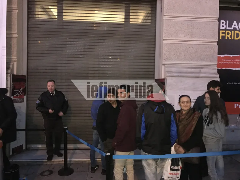 Άνθρωποι περιμένουν σε ουρά έξω από κατάστημα με κλειστά ρολά για την Black Friday.