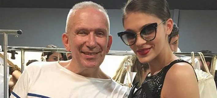 Ο Jean Paul Gaultier χαμογελά δίπλα σε μοντέλο με γυαλιά ηλίου στα παρασκήνια επίδειξης μόδας.