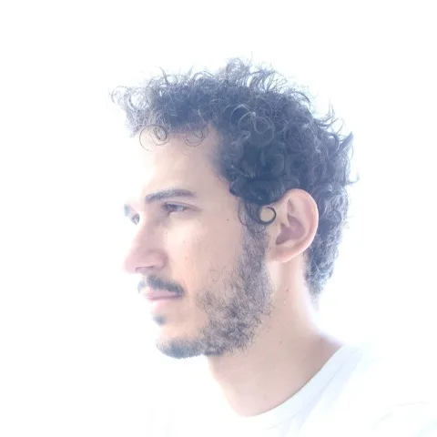 Προφίλ του καλλιτέχνη Lumiere με σγουρά μαλλιά και μούσι σε λευκό φόντο.