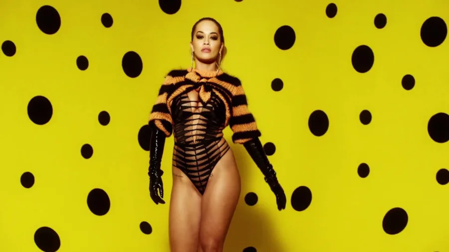 Η Rita Ora με μαύρο κορμάκι και ριγέ μπολερό σε κίτρινο φόντο με βούλες.