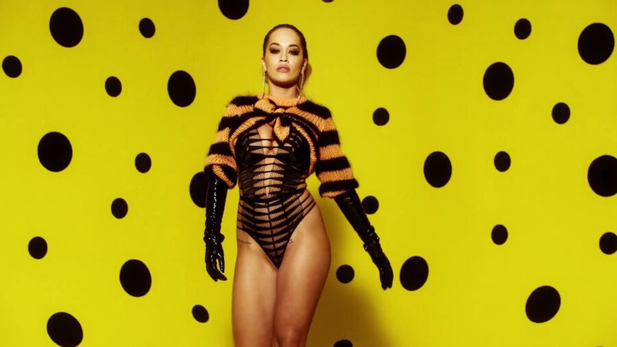 Η Rita Ora με μαύρο κορμάκι και ριγέ μπολερό σε κίτρινο φόντο με βούλες.