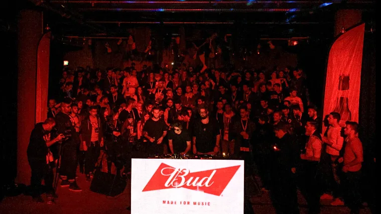 Πλήθος κόσμου σε εκδήλωση Bud Made For Music με DJ και κόκκινο φωτισμό.