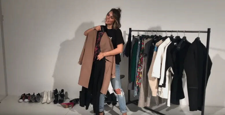 Η Elena Galifa κρατάει ένα καμηλό παλτό μπροστά από μια κρεμάστρα με ρούχα.