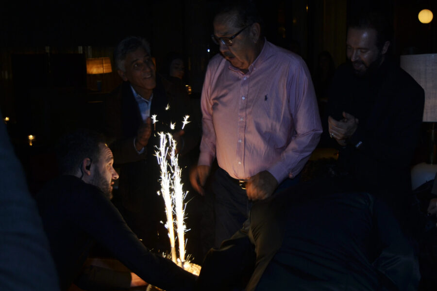 Άνδρες γύρω από μια βασιλόπιτα με αναμμένα πυροτεχνήματα σε σκοτεινό χώρο.