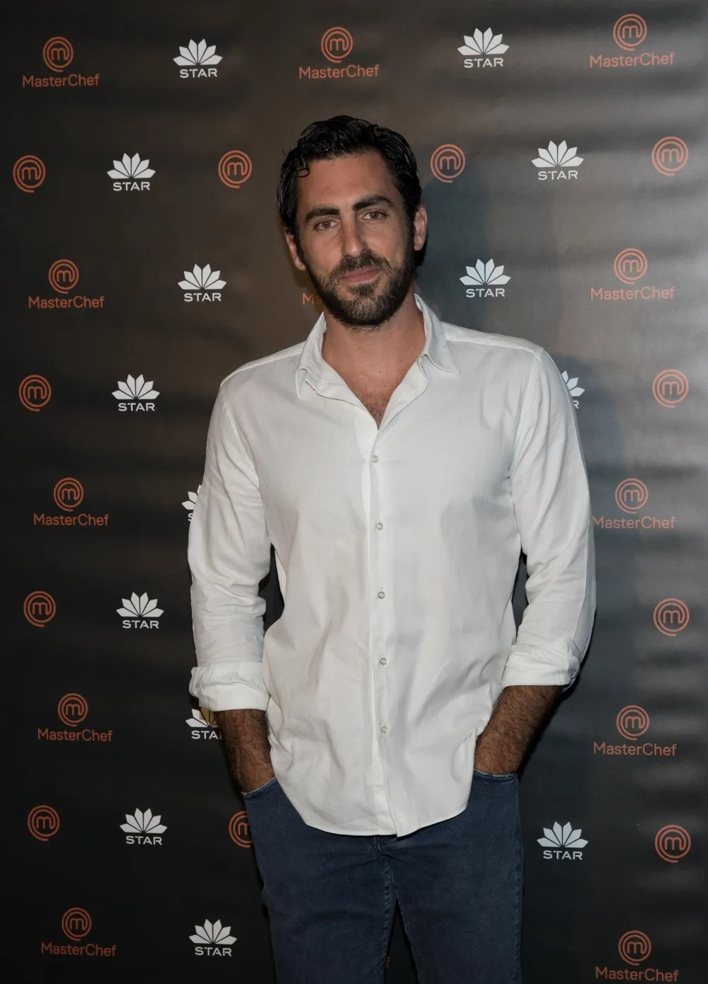 Ο σεφ του MasterChef 2, Λεωνίδας Κουτσόπουλος, ποζάρει μπροστά στο backdrop του διαγωνισμού μαγειρικής.