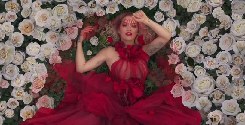Η Rita Ora ξαπλωμένη ανάμεσα σε λευκά τριαντάφυλλα φορώντας κόκκινο φόρεμα.