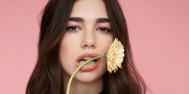 Η Dua Lipa κρατάει ένα κίτρινο λουλούδι στο στόμα της σε ροζ φόντο.