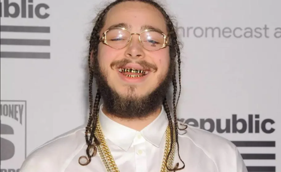 Ο Post Malone χαμογελάει φορώντας γυαλιά και χρυσά δόντια.