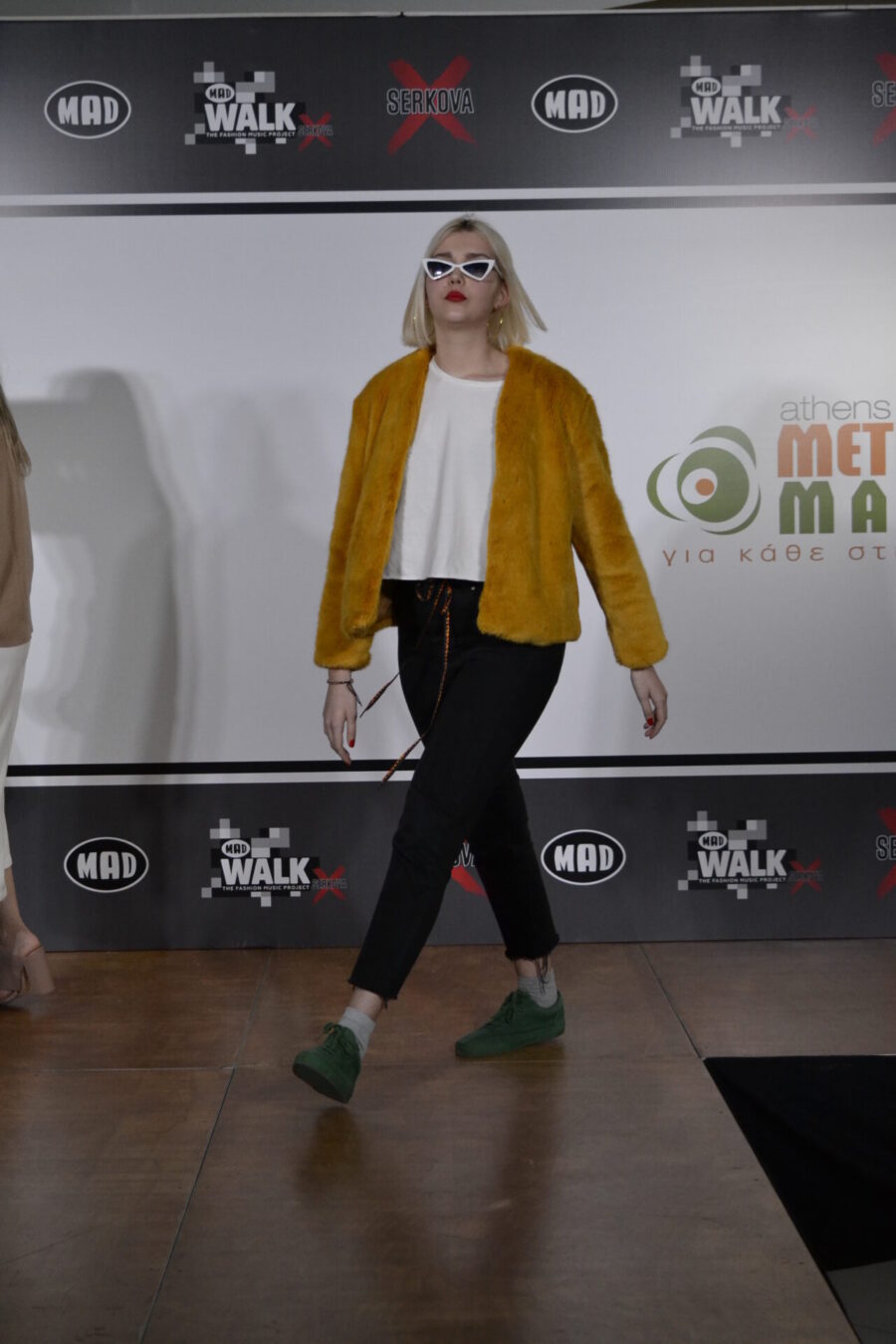 Καλλιτέχνης με κίτρινη γούνα και λευκά γυαλιά ηλίου σε πασαρέλα.