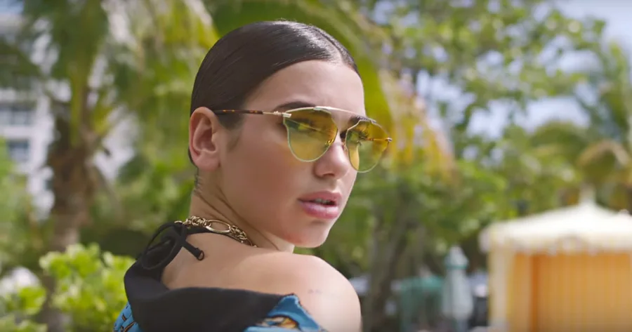 Η Dua Lipa με κίτρινα γυαλιά ηλίου και πιασμένα μαλλιά σε εξωτερικό χώρο.