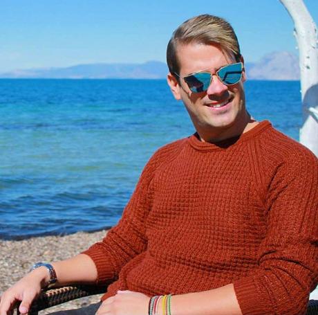 Άνδρας με καφέ πλεκτό πουλόβερ και γυαλιά ηλίου καθισμένος δίπλα στη θάλασσα.