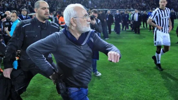 Άνδρας με γκρίζα μαλλιά και γυαλιά σε γήπεδο ποδοσφαίρου κατά τη διάρκεια έντασης.