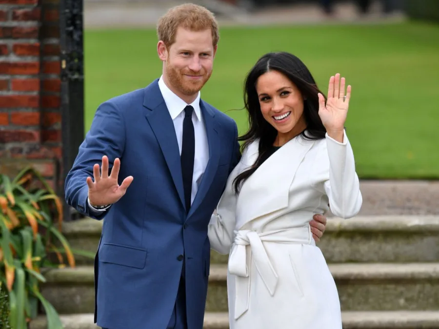 Ο Prince Harry και η Meghan Markle χαιρετούν χαμογελαστοί σε εξωτερικό χώρο.