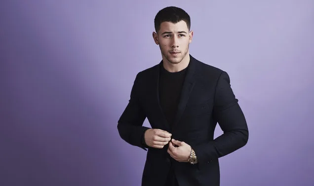 Ο Nick Jonas ποζάρει με μαύρο κοστούμι σε μωβ φόντο.
