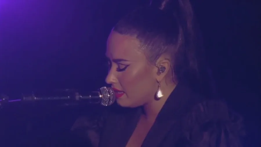 Η Demi Lovato τραγουδάει σε μικρόφωνο σε σκοτεινή σκηνή με μωβ φωτισμό.